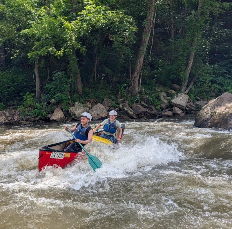 Two young people are paddling a red canoe through some rapids. They are both wearing helmets and life jackets. The person in the front is paddling with a blue paddle, and the person in the back is also paddling. The water is choppy and white, and there are trees and rocks in the background.
